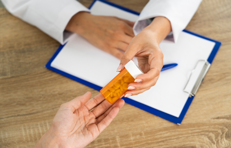 Healthcare professional handing an orange prescription pill bottle to a patient across a desk, with a clipboard and pen in the background