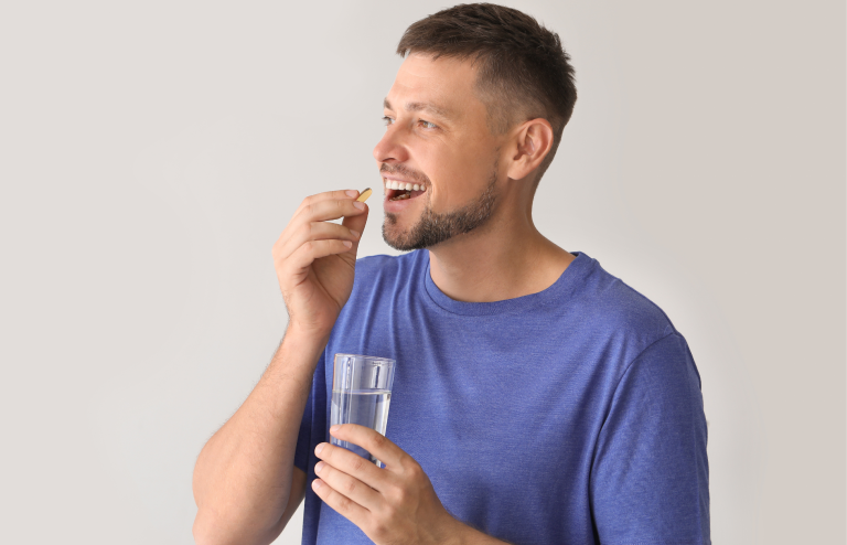 Smiling adult man in a blue T-shirt holding a glass of water while placing a pill into his mouth against a neutral background