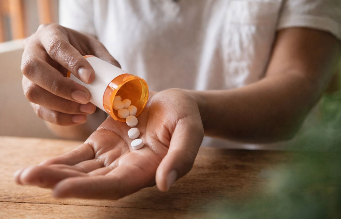 Close-up of a person’s hands pouring white oral medication pills from an orange prescription bottle into their open palm on a wooden table.