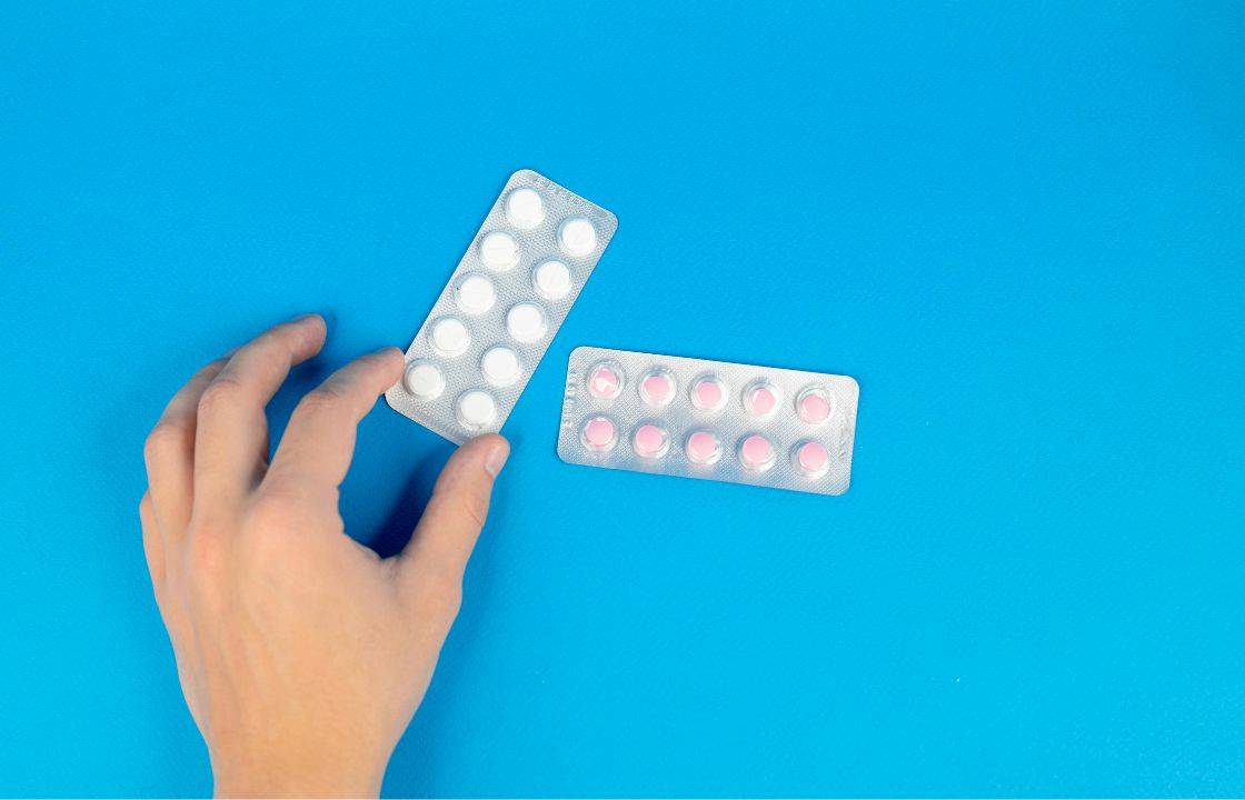Hand holding a blister pack of white GLP-1 pills next to another blister strip of pink tablets on a bright blue background.