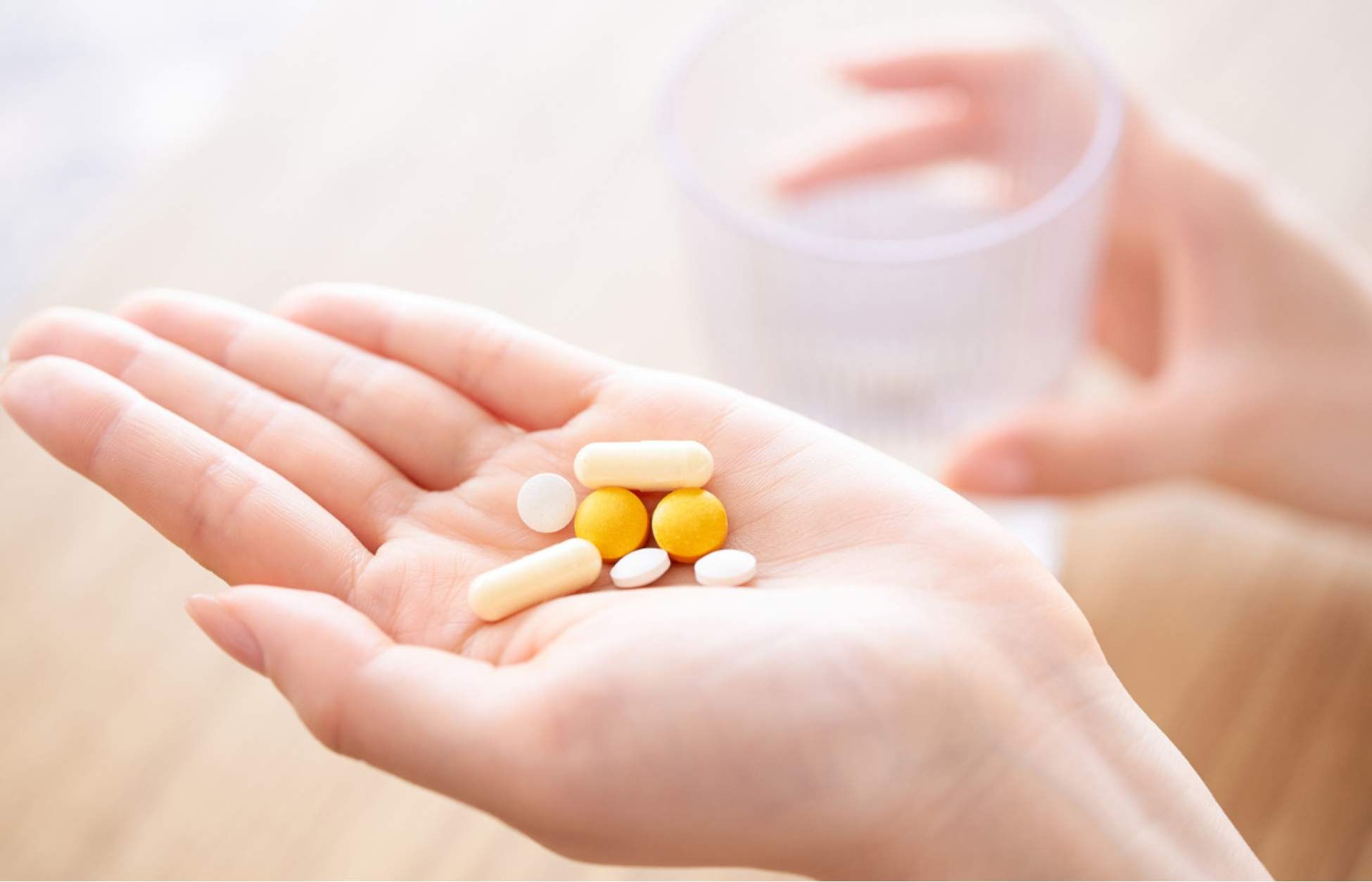 Hand holding assorted pills and capsules with a glass of water in the background.