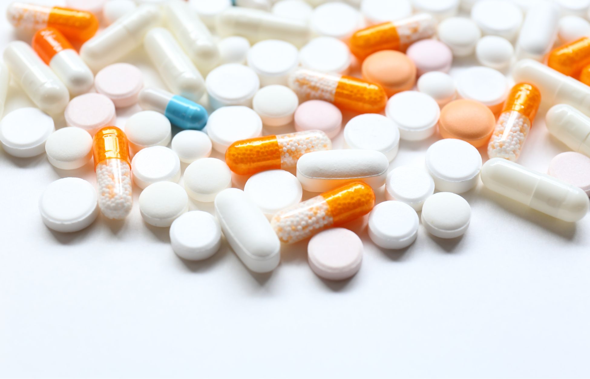 Assortment of white tablets, pink round pills, and orange-and-clear capsules scattered on a white surface.