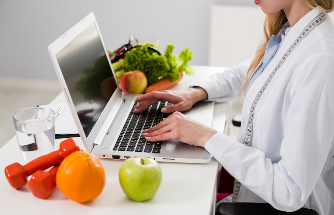 A healthcare professional using a laptop at a desk with fresh fruits, vegetables, a glass of water, and dumbbells, representing digital nutrition and weight-management care.