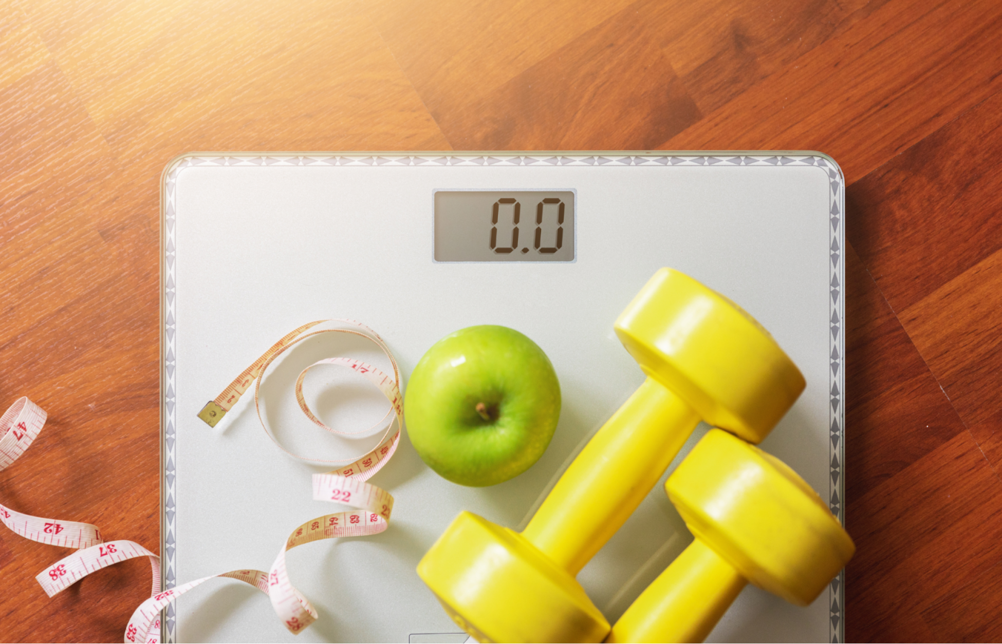 Bathroom scale with a green apple, yellow dumbbells, and a measuring tape on a wooden floor.
