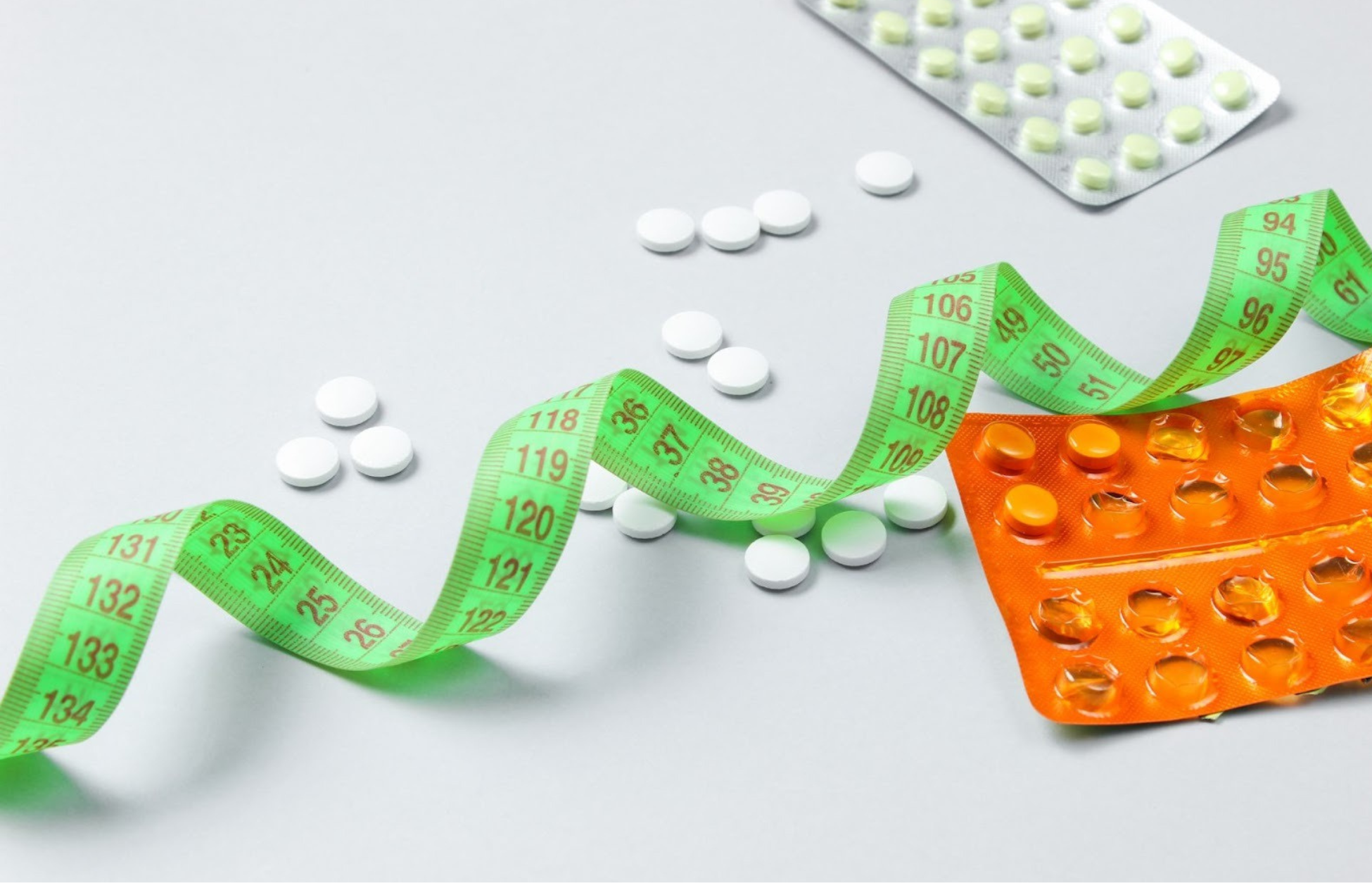 White tablets scattered beside an orange blister pack and a green measuring tape on a light background.