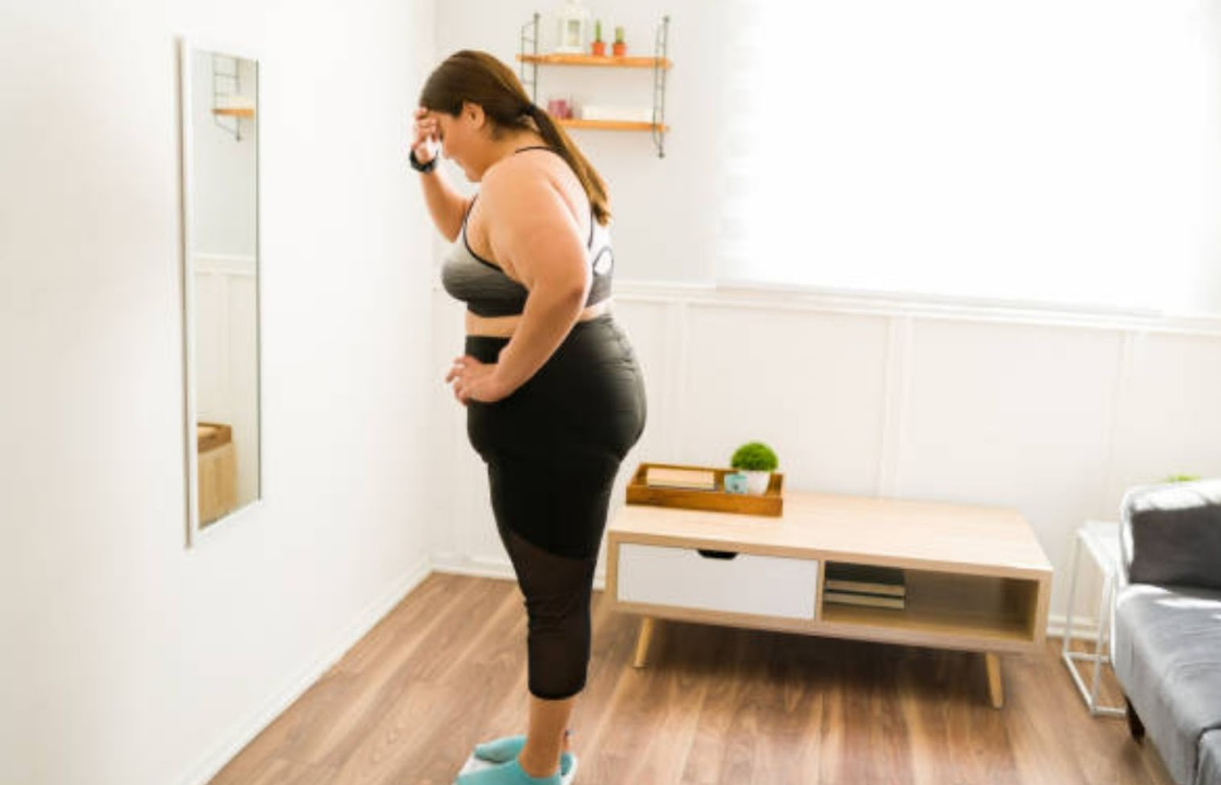 Woman in workout clothes standing on a scale in a bright living room, looking concerned while touching her forehead.