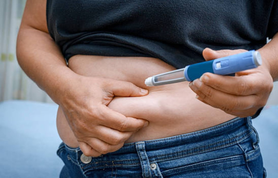 Person holding a GLP-1 injection pen near their abdomen while pinching belly fat, preparing for a weight-loss or diabetes medication shot.