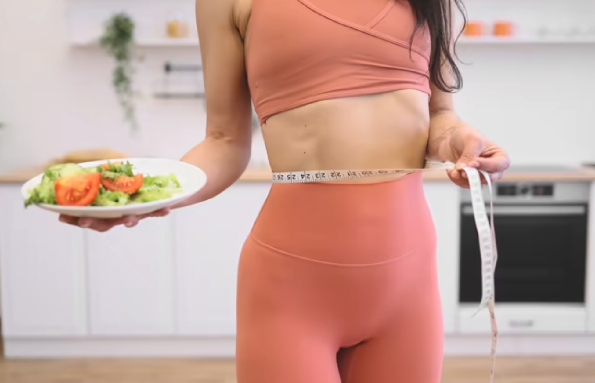 Person measuring waist while holding a plate of salad in a kitchen.