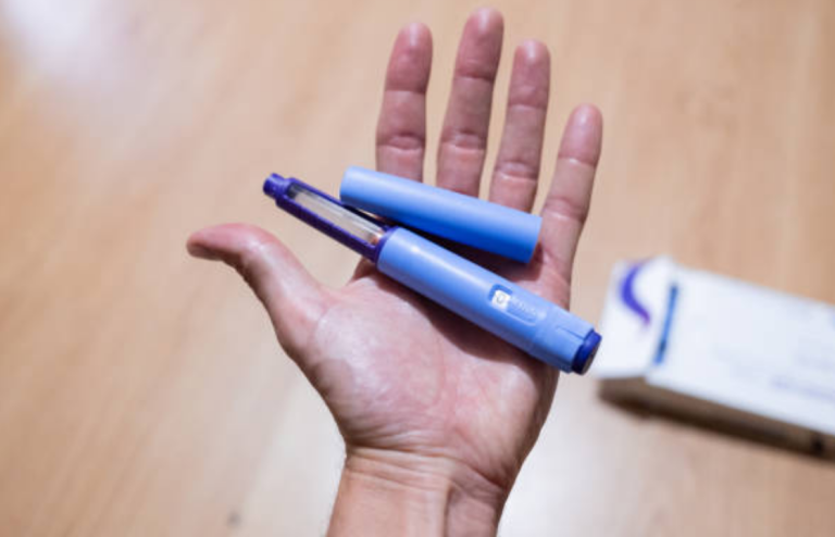 Hand holding a blue injectable medication pen with its cap removed, likely a GLP-1 device.