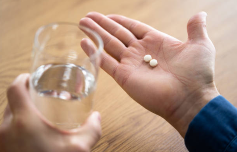 Hand holding two pills with a glass of water nearby.