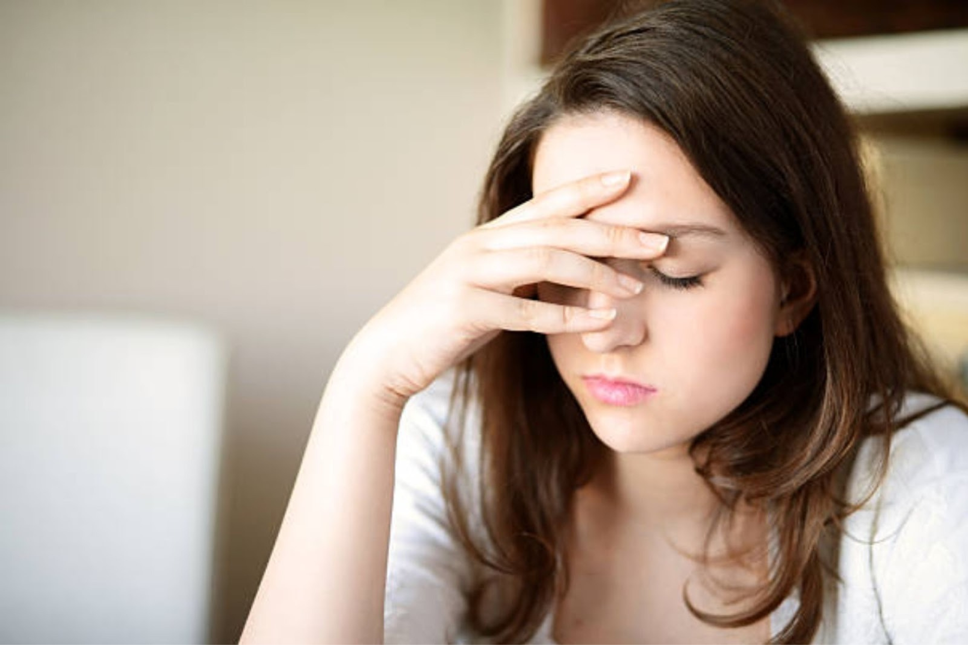 A woman sitting indoors with her hand on her forehead, appearing stressed or experiencing a headache.