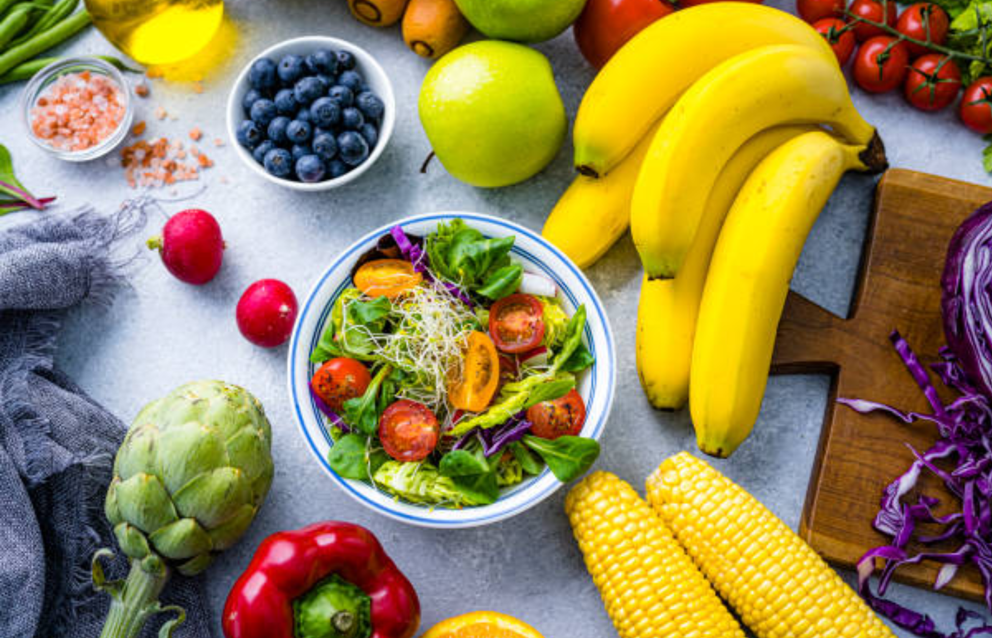Colorful assortment of fresh fruits and vegetables with a bowl of salad, bananas, corn, blueberries, and leafy greens.