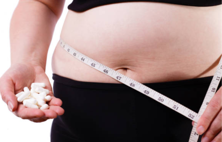 Person measuring waist with a tape measure while holding white pills in one hand.