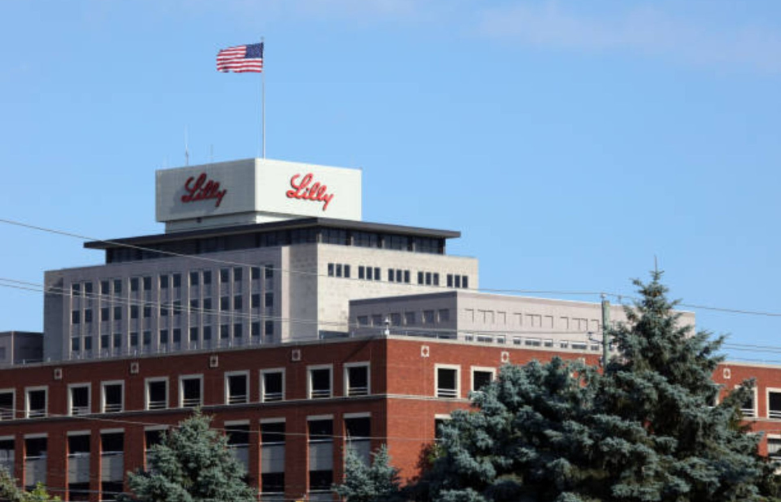 Eli Lilly corporate headquarters building with the company logo displayed on the rooftop tower and an American flag flying above the campus on a clear day.
