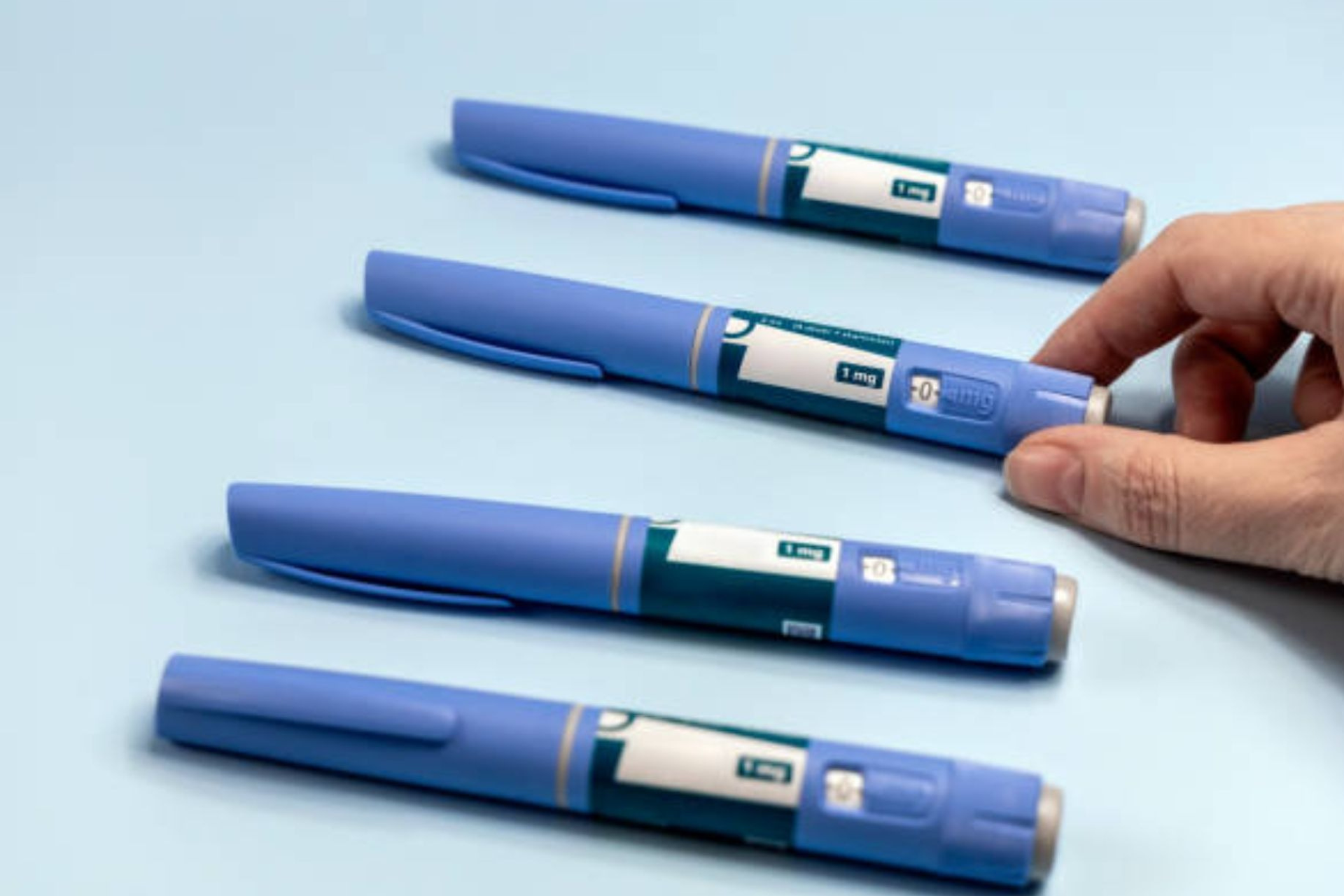 Blue GLP-1 injection pens placed on a light blue surface with a hand reaching toward one pen.