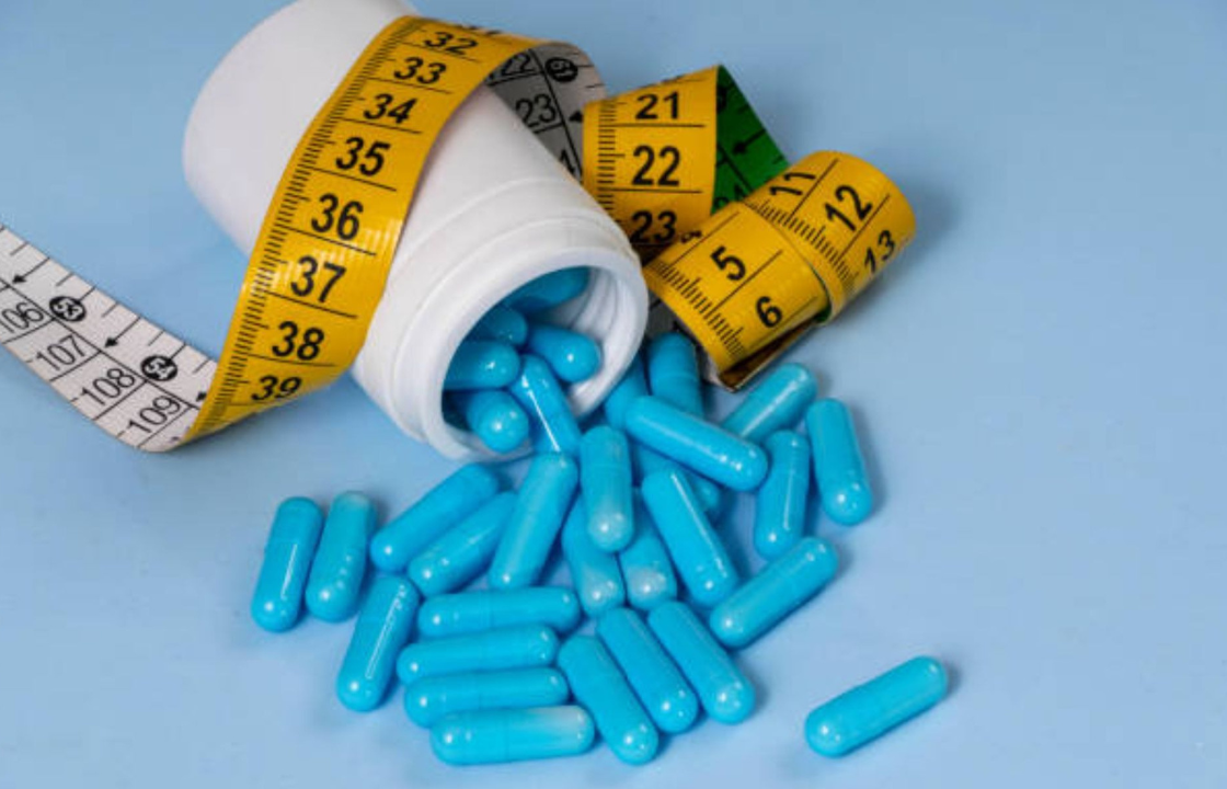 Blue weight-loss capsules spilling from a white pill bottle beside measuring tapes on a light blue background.