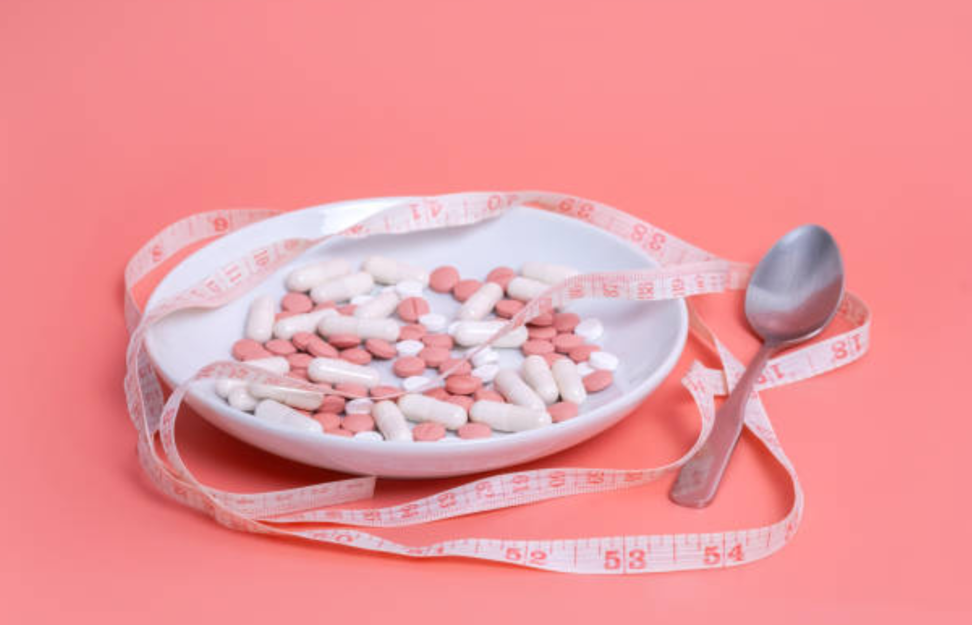 Plate filled with weight loss pills and capsules surrounded by a measuring tape on a pink background.