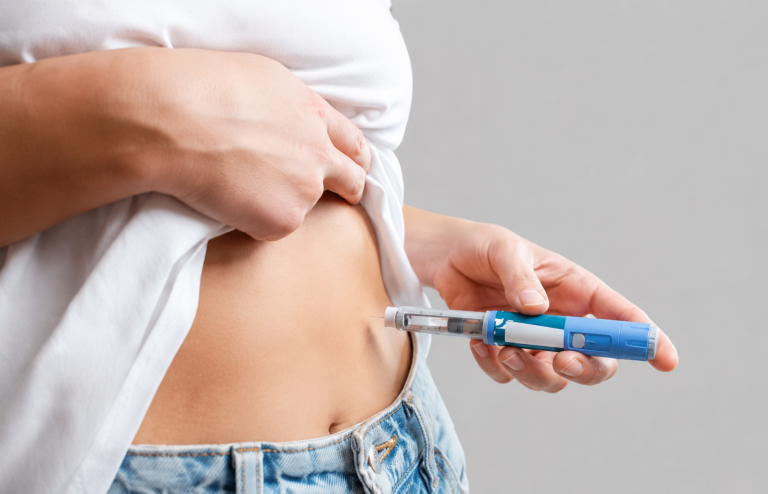 Person using a blue injection pen to administer a shot into the abdomen against a light grey background.