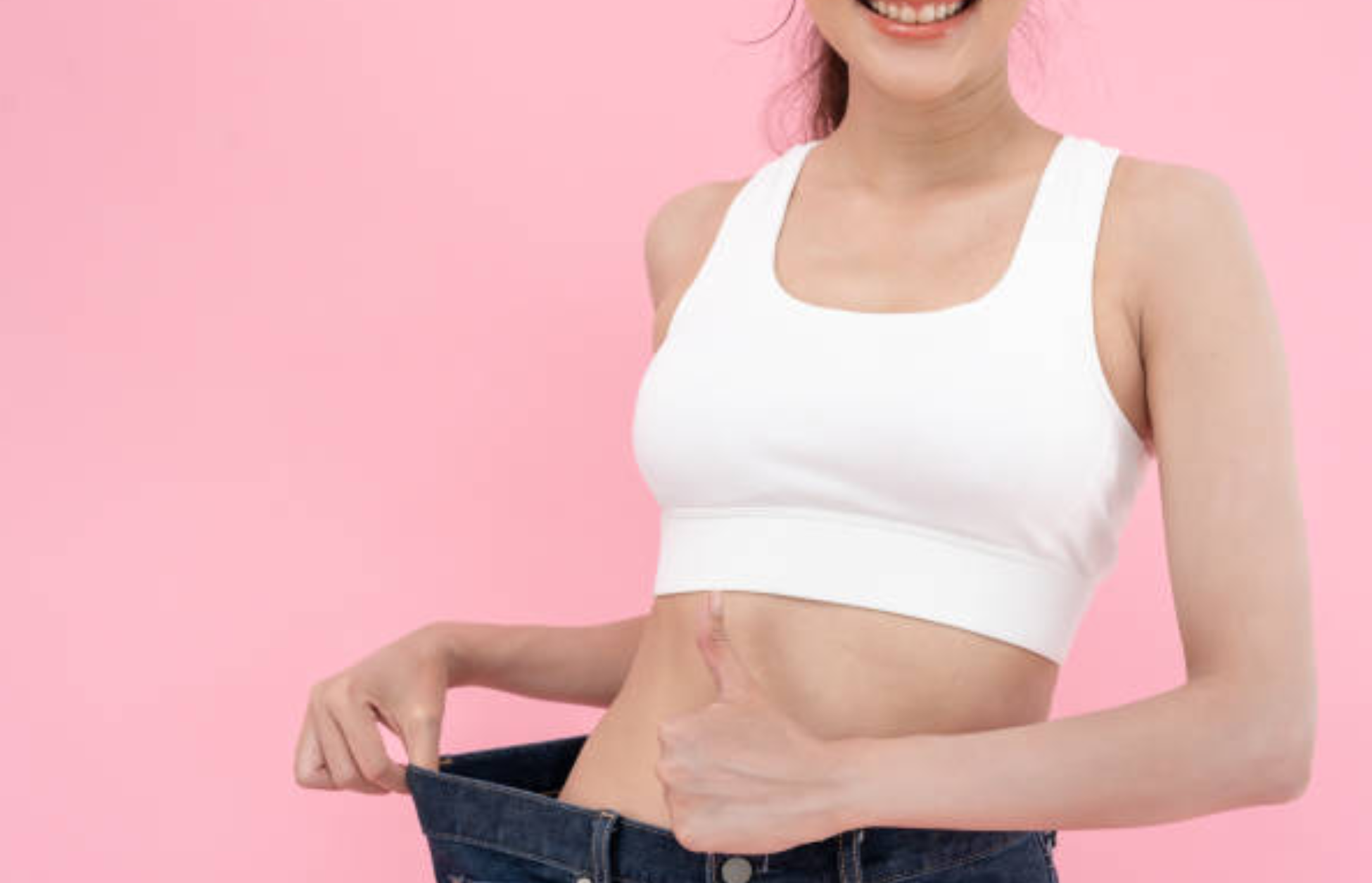 Woman showing weight loss by holding loose-fitting jeans against a pink background