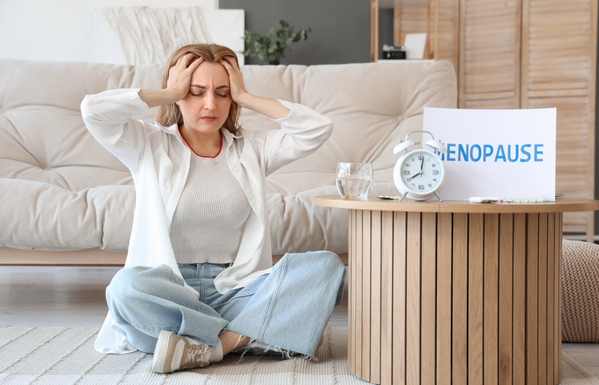 Woman sitting on the floor holding her head beside a table with a clock, water glass, pills, and a sign reading “Menopause.”