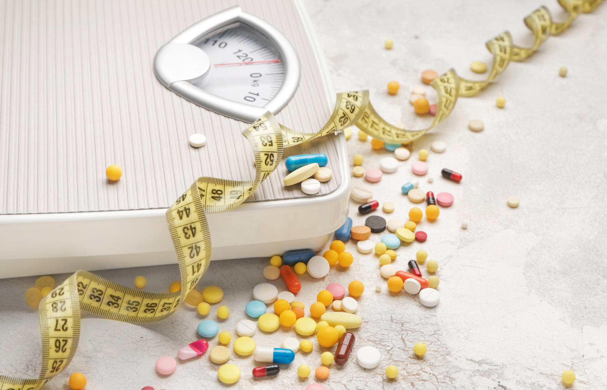 Bathroom scale with a measuring tape and colorful pills scattered around, representing weight-loss medications and obesity treatment.