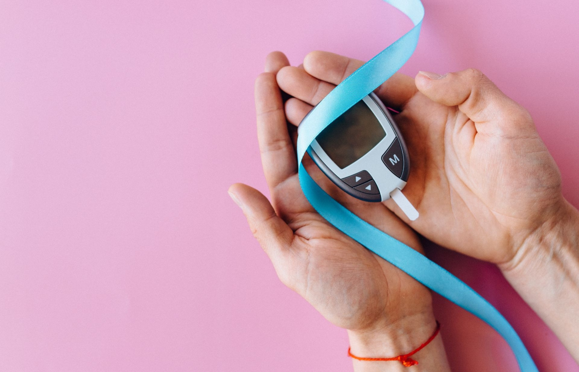 Hands holding a blood glucose meter wrapped with a blue awareness ribbon on a pink background.