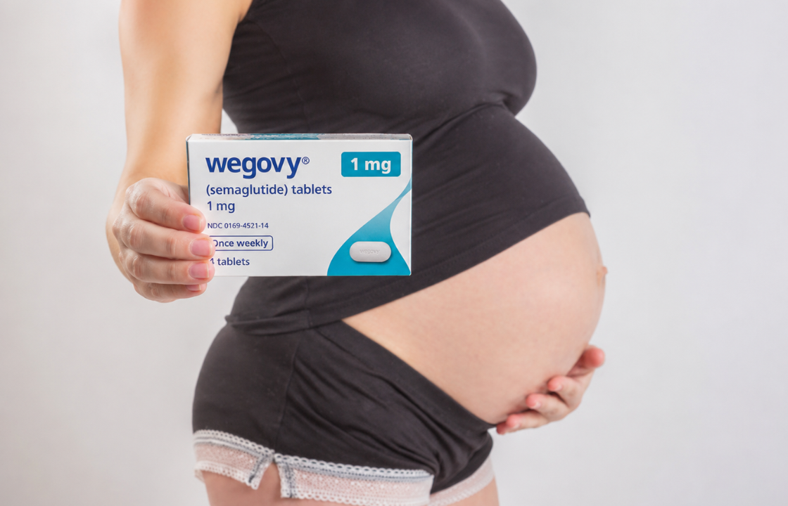 Pregnant woman holding a Wegovy medication box toward the camera while gently supporting her baby bump with her other hand, standing against a light gray studio background.