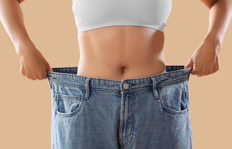 Close-up of a person wearing a white sports bra and holding out oversized blue jeans to show significant weight loss.