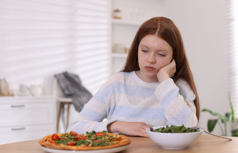 Young woman looking thoughtfully at a salad and pizza on a dining table.