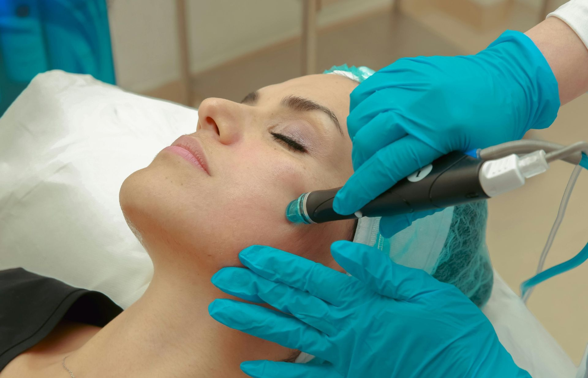 A patient receives a noninvasive facial skin treatment while a clinician uses a handheld cosmetic device and wears blue gloves.