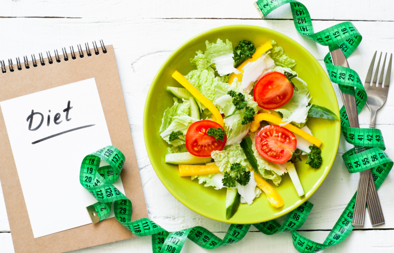 Fresh vegetable salad with lettuce, tomatoes, cucumbers, and bell peppers beside a diet notebook and measuring tape.