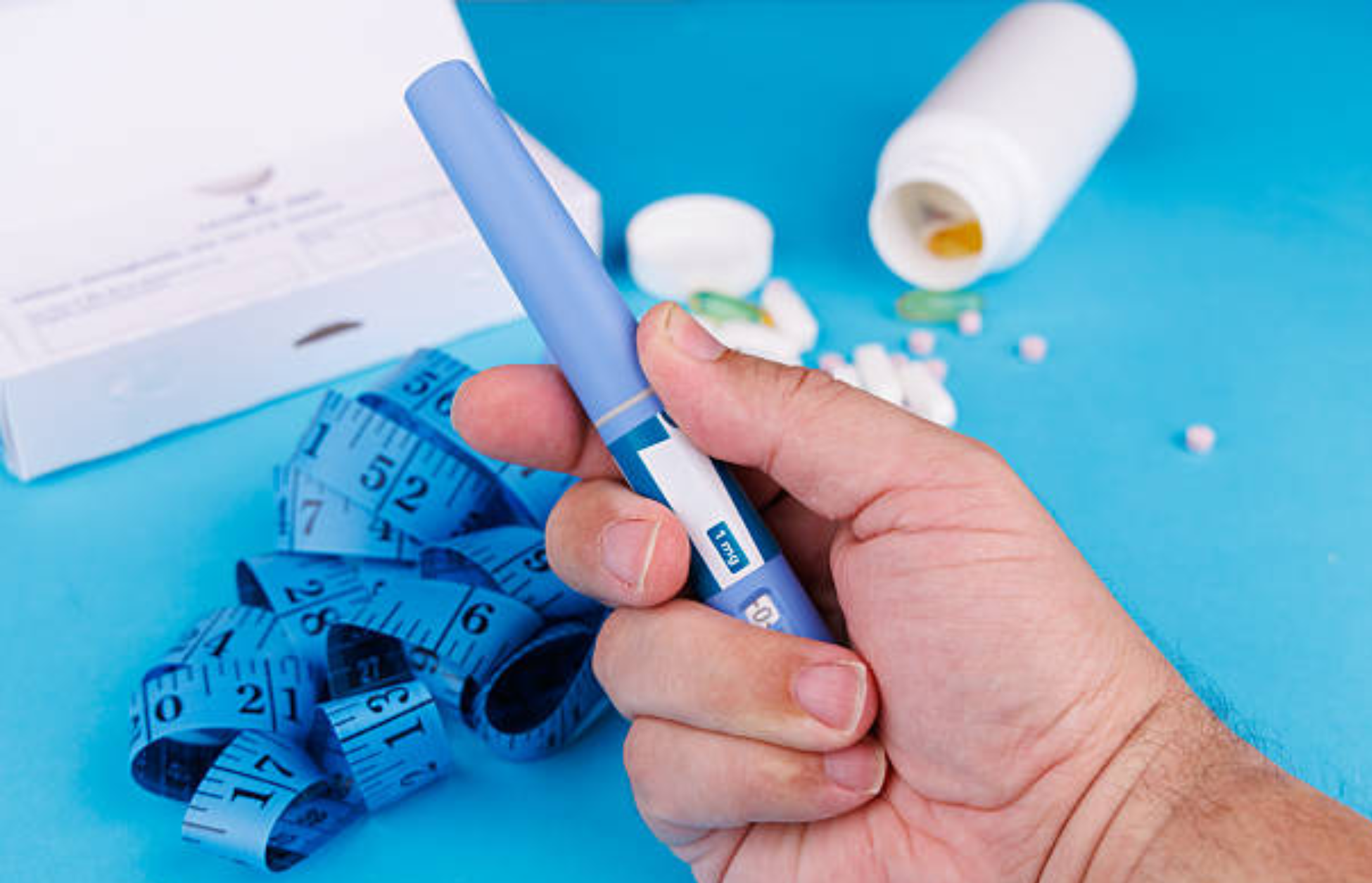 Hand holding a blue GLP-1 injection pen beside a measuring tape and medication bottles on a blue surface.