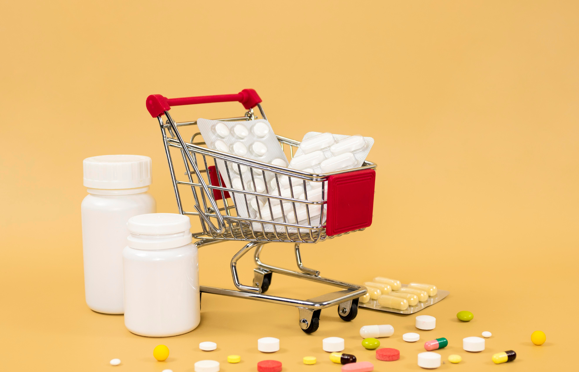 Mini shopping cart filled with blister packs of pills beside medicine bottles and scattered tablets on a yellow background.