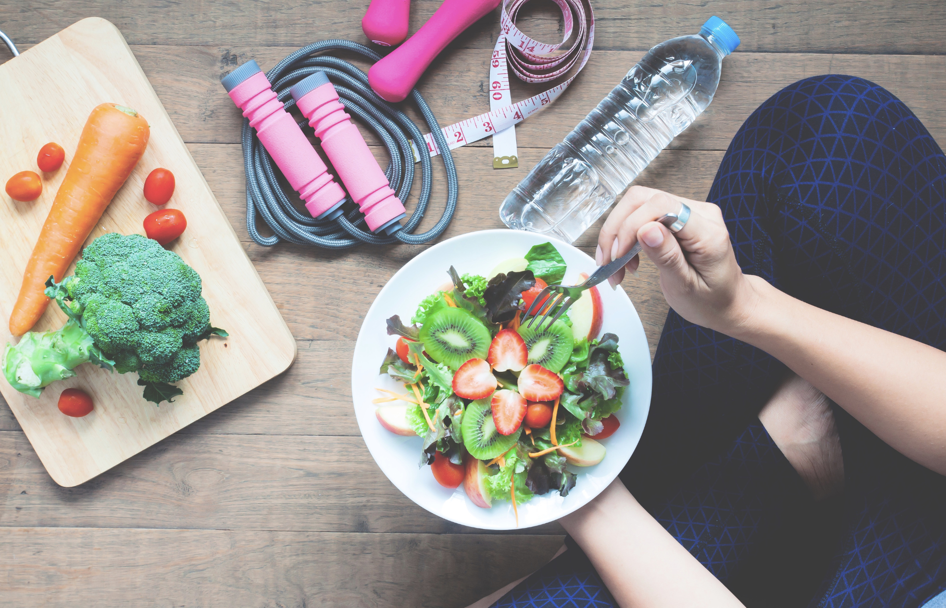 Healthy salad, fresh vegetables, water bottle, and fitness equipment arranged beside a person eating a nutritious meal.