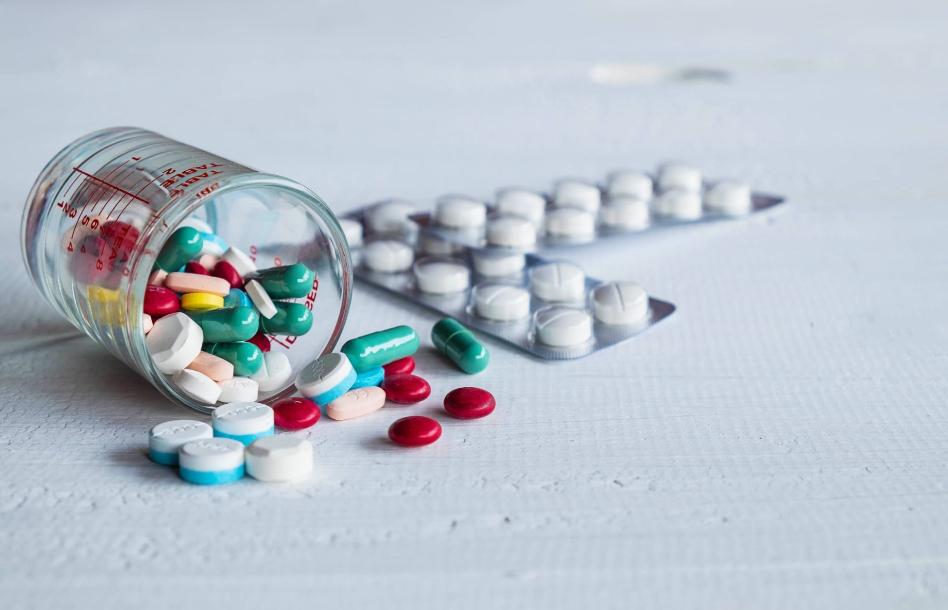 Assorted colorful pills and capsules spilled from a medicine cup beside a blister pack of tablets on a white surface.