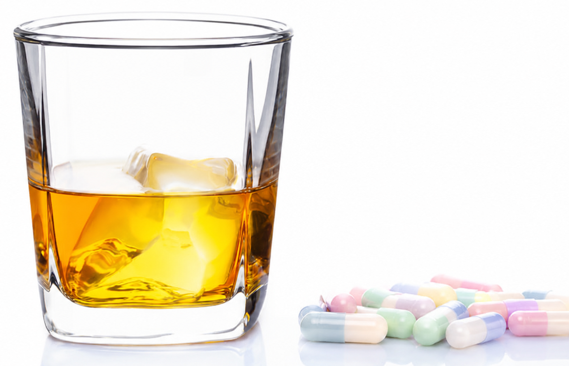 Alt Text: A glass of amber whiskey with ice cubes sits next to a small pile of light-colored capsules on a white background.