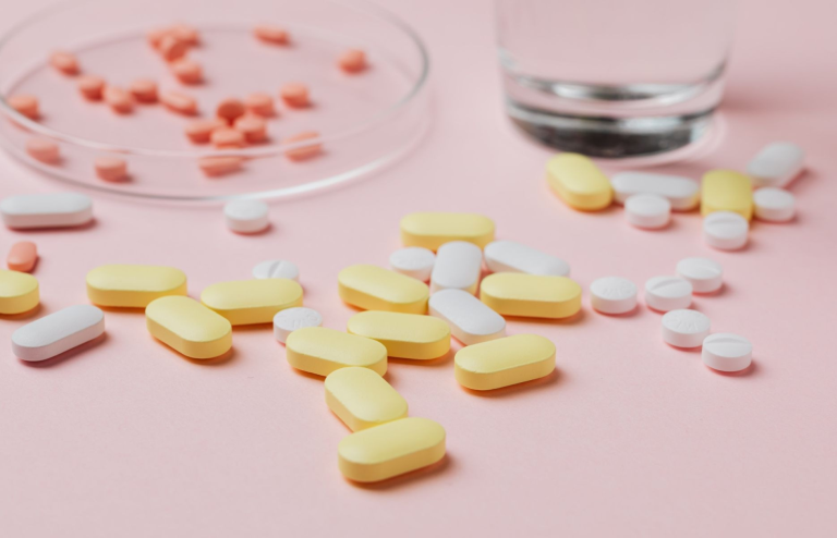 Yellow and white pharmaceutical tablets scattered on a pink surface beside a glass of water.
