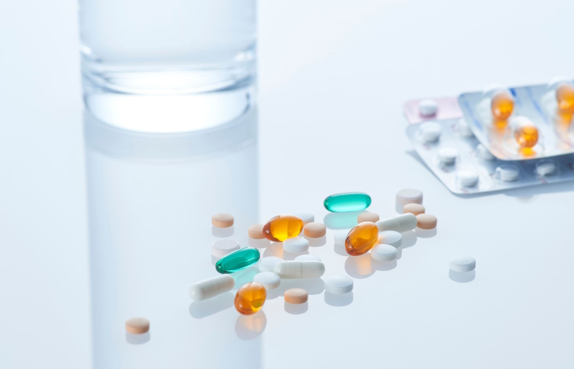 Assorted pills and capsules scattered on a white surface beside a glass of water and blister packs, representing oral medication use.