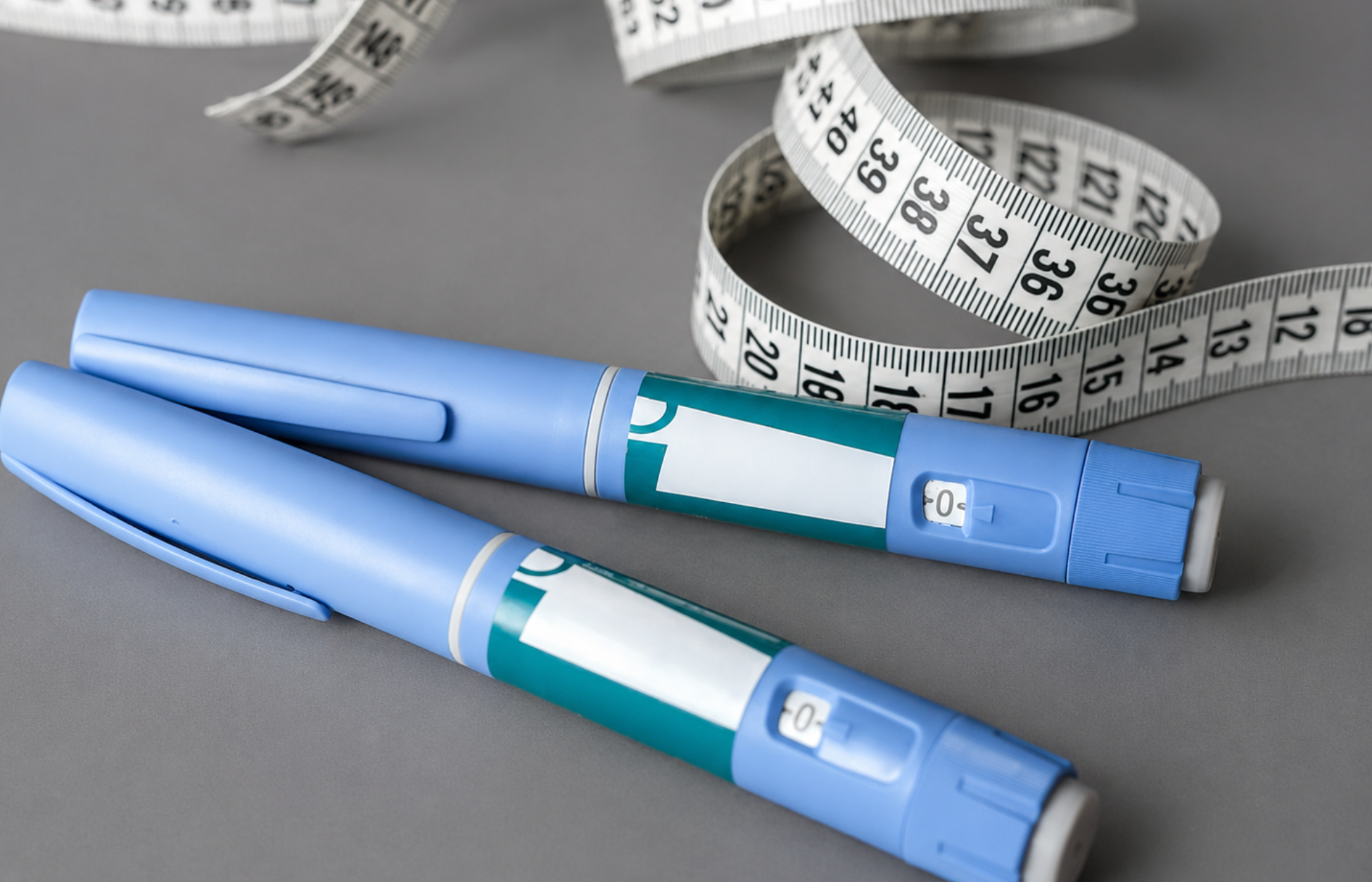 Two blue injection pens beside a measuring tape on a gray background, symbolizing weight loss medication.