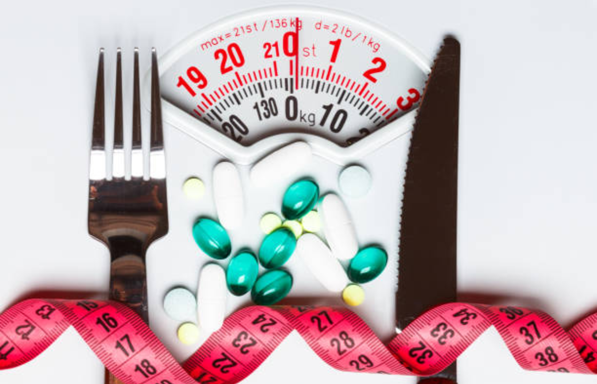 Weight scale filled with pills, alongside a fork, knife, and measuring tape, symbolizing medical weight-loss treatment.