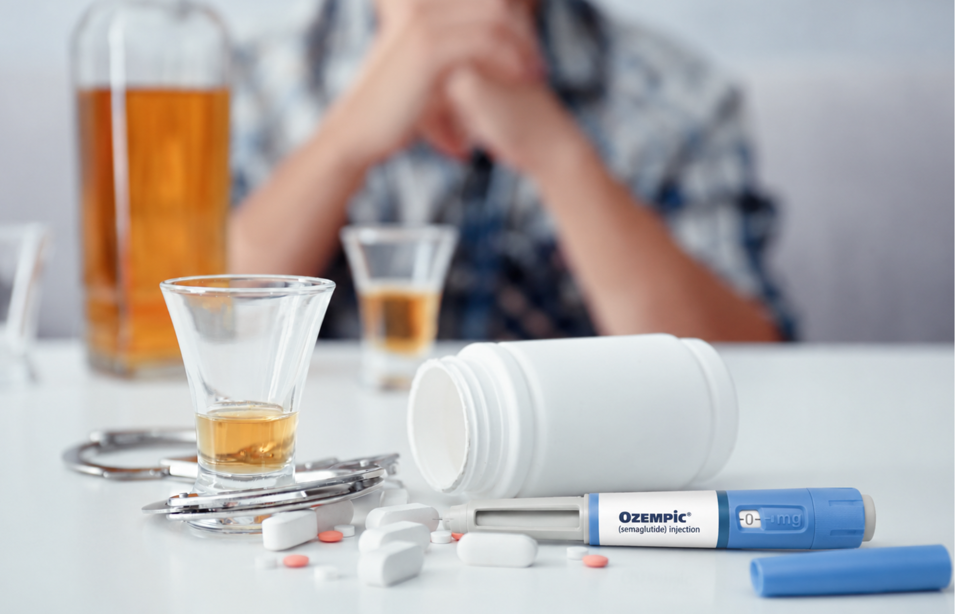 GLP-1 injection pen placed beside alcohol shots, pills, and an overturned pill bottle on a table, with a blurred person sitting in the background.