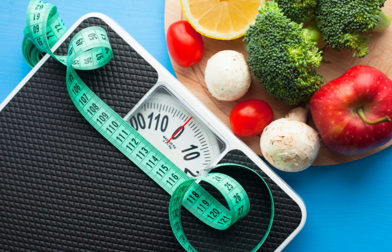 Bathroom scale with a measuring tape beside fresh fruits and vegetables, representing healthy weight management and nutrition.