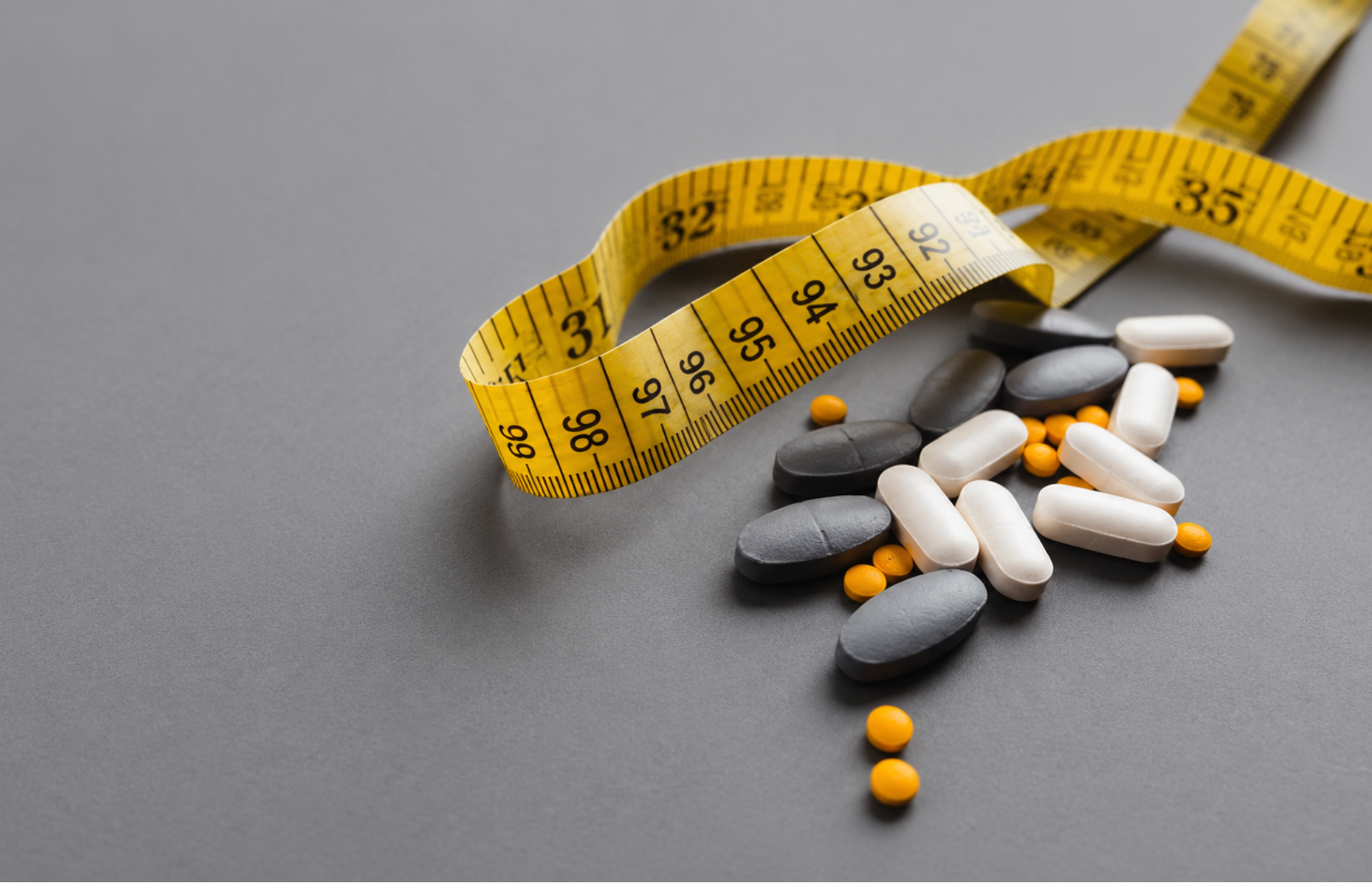 Yellow measuring tape wrapped around a pile of white, gray, and orange pills on a smooth gray background.