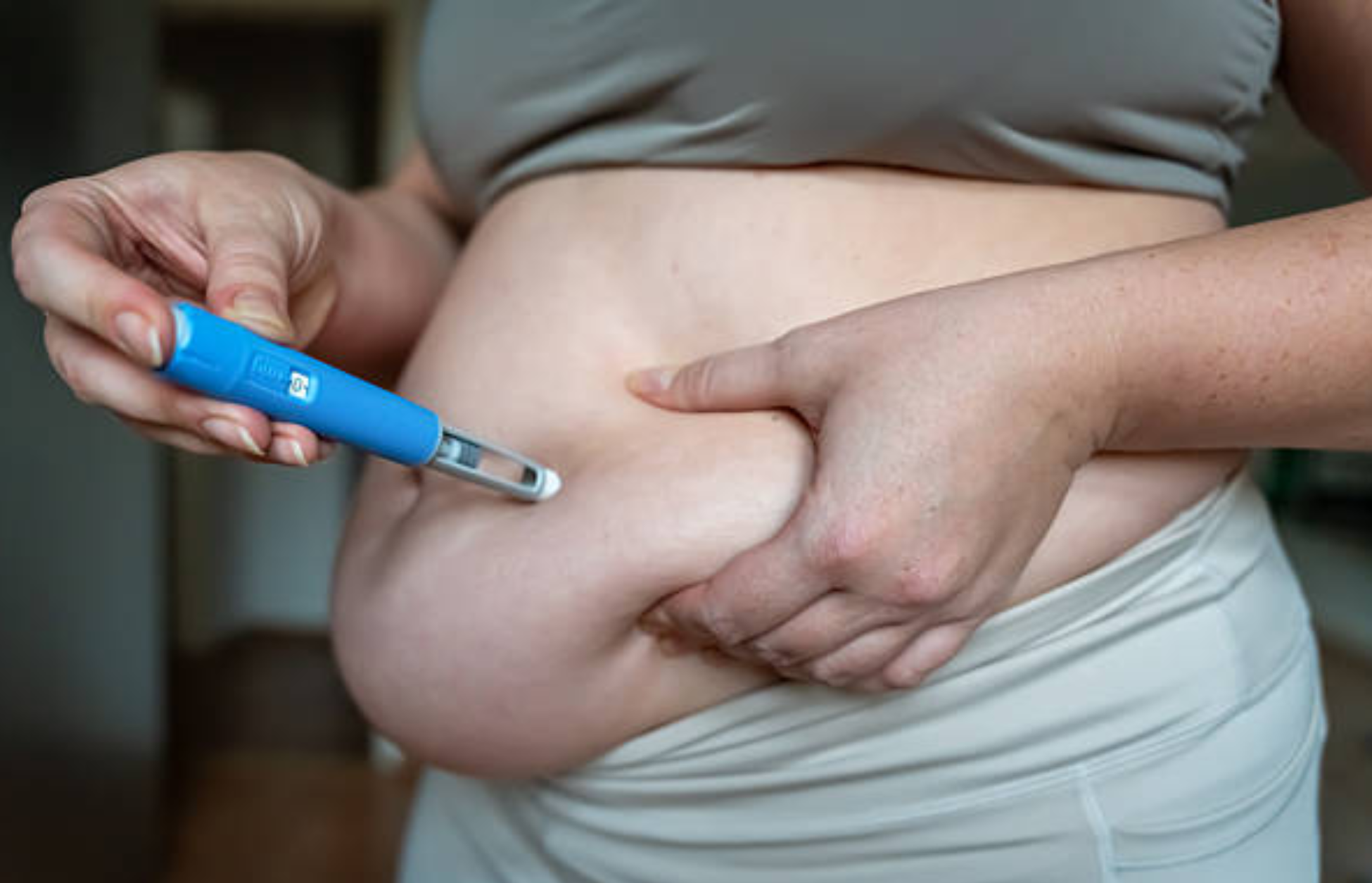 Person pinching abdominal fat while holding a blue injection pen against their stomach, illustrating a weight-loss medication injection.